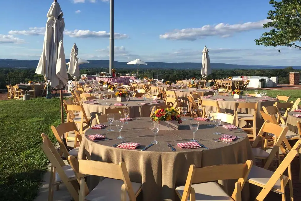 Image of an outdoor event setup with round tables covered in beige tablecloths and surrounded by wooden chairs. Tables feature floral centerpieces and place settings, with umbrellas providing shade.