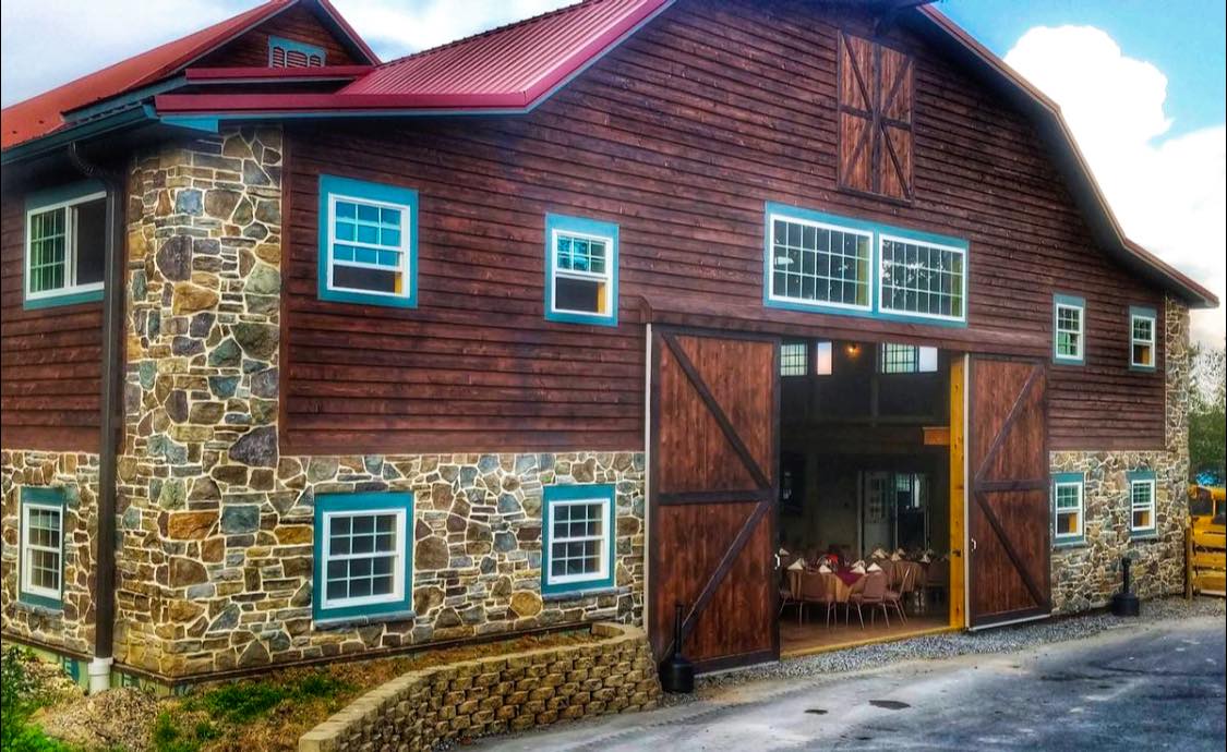 A rustic barn with a stone and wood exterior, featuring large open double doors, provides the perfect backdrop for a charming Gettysburg wedding expo. Inside, tables and chairs are set up, ready to welcome guests to this enchanting event.