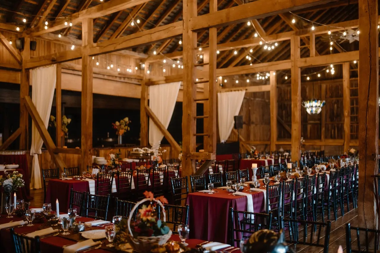 Rustic indoor wedding reception with wooden beams and string lights, reminiscent of a Gettysburg Wedding Expo. Tables are set with burgundy tablecloths, floral centerpieces, and chairs in rows, creating an enchanting atmosphere for your special day.