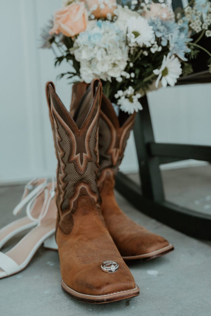 Brown cowboy boots with a wedding bouquet inside and wedding rings on the toe. A pair of white heels behind them.