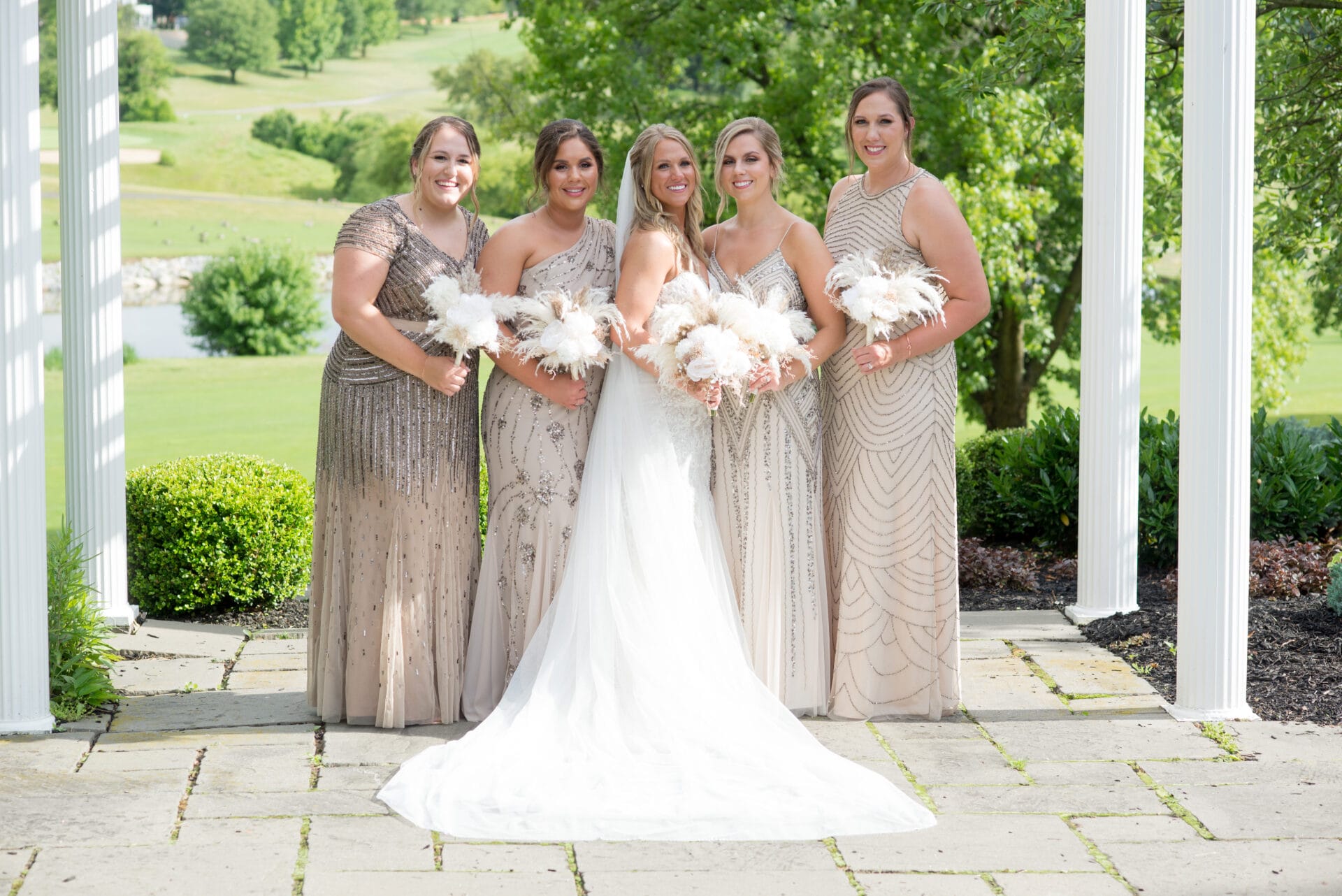 Bride in white wedding dress standing with four bridesmaids in different taupe-colored dresses holding white flower bouquets.