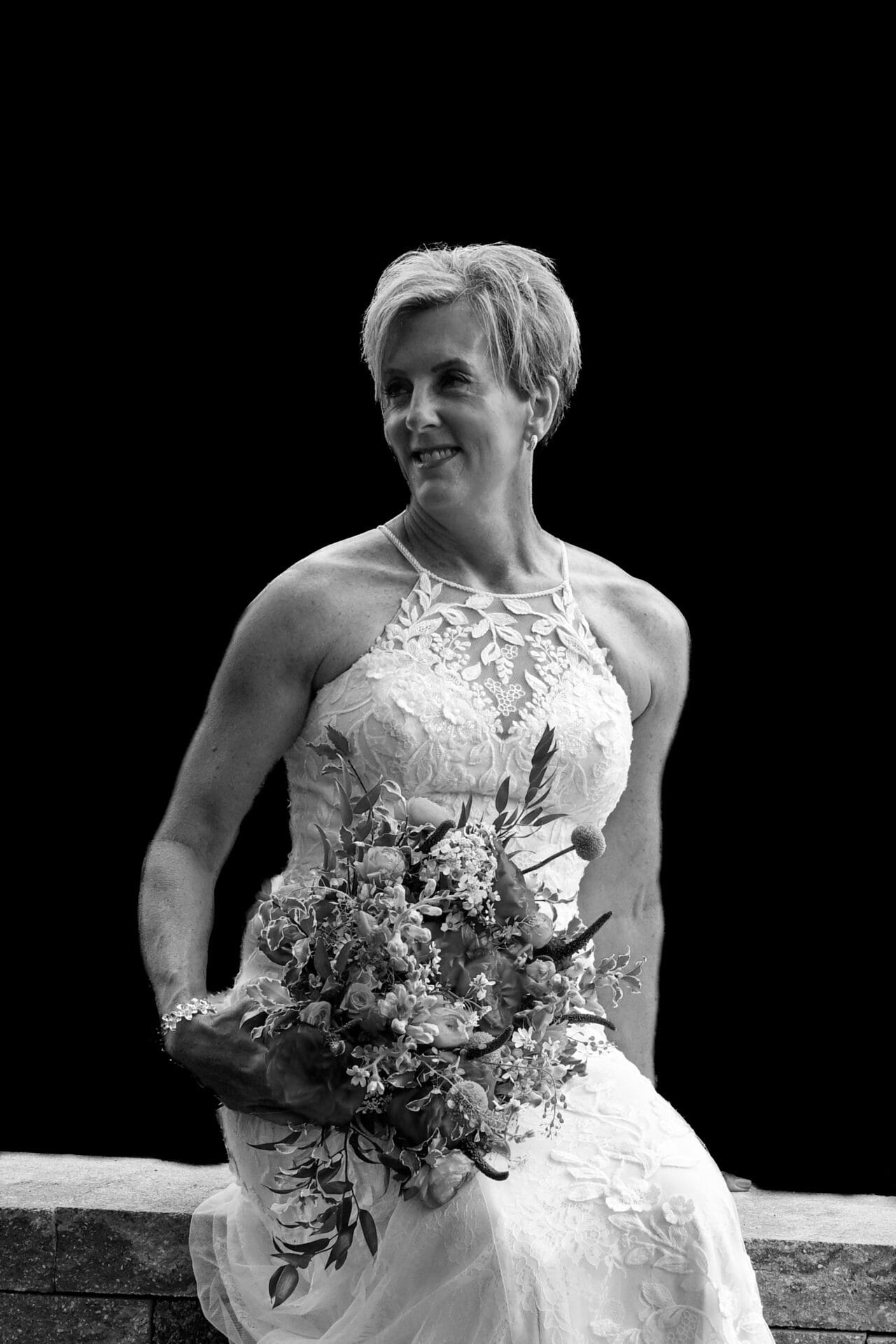 Bride wearing lace halter neck wedding dress holding flower bouquet in black and white.