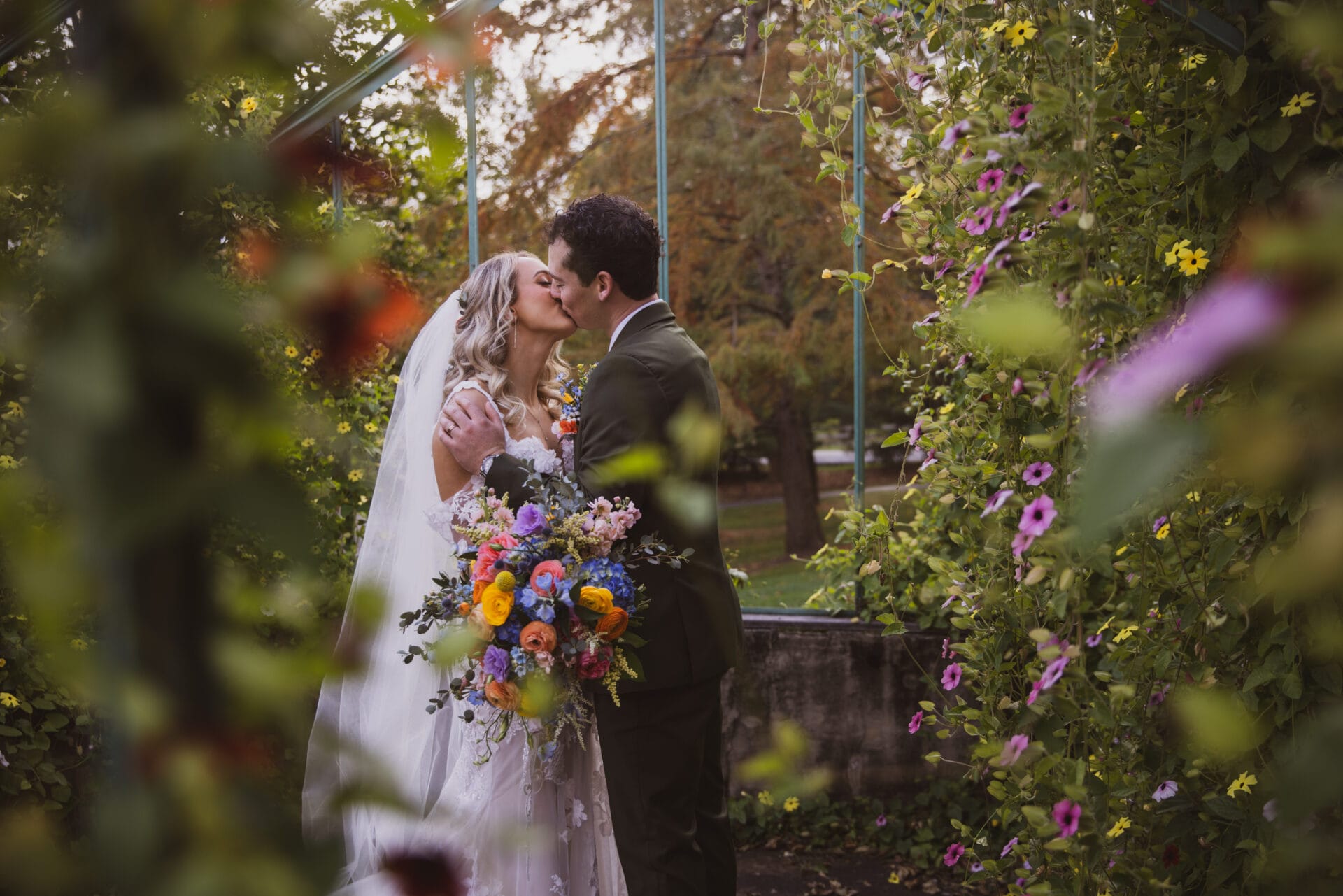 Couple kissing in garden on their wedding day surrounded by flowers and foliage.